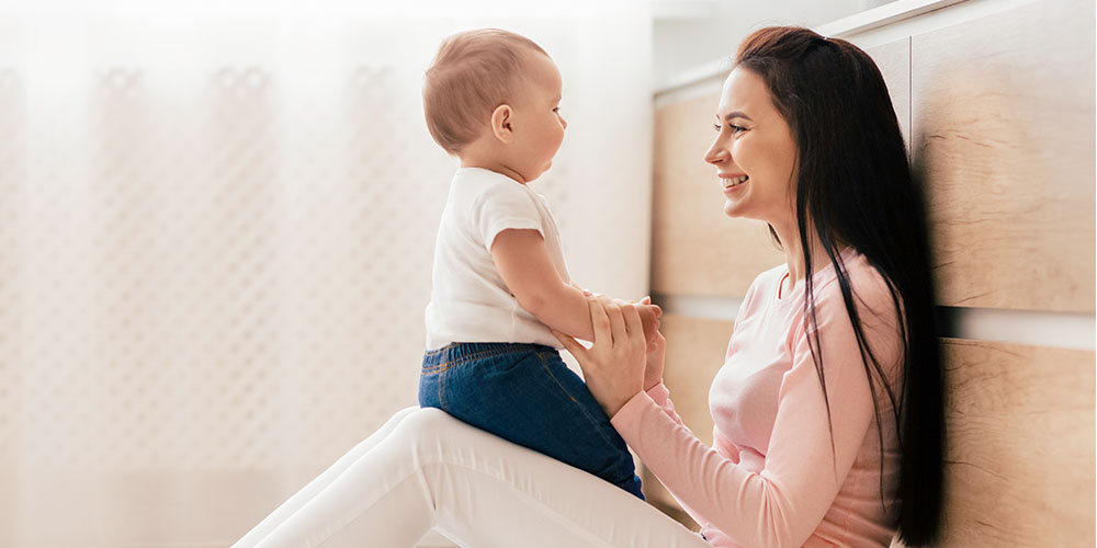 Baby sitzt auf den Beinen der Mutter, während beide sich lachend anschauen.