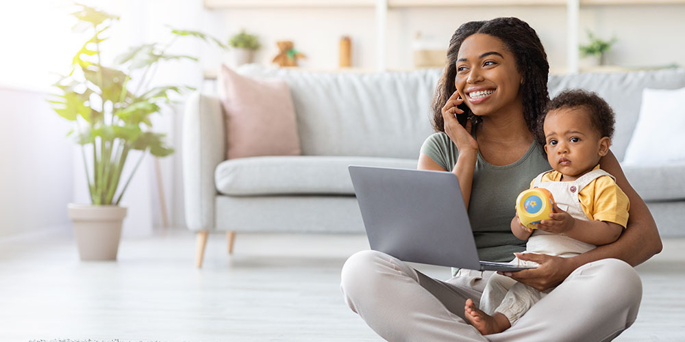 Junge Mutter telefoniert und arbeitet während der Elternzeit am Laptop, während das Baby auf ihrem Schoß sitzt. Sie nutzt den Zuverdienst beim Elterngeld.