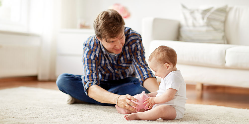 Vater sitzt mit seinem Baby im Wohnzimmer auf dem Boden und zeigt ihm eine Spardose.