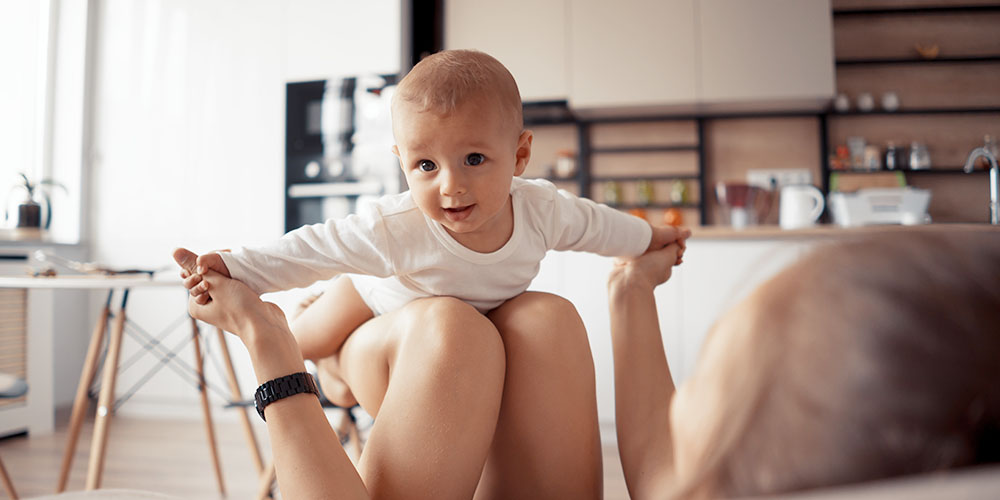 Baby liegt auf dem Bauch auf den Beinen der Mutter und macht eine fliegende Bewegung, während die Mutter seine Hände hält. Im Hintergrund ist eine Küche zu sehen.