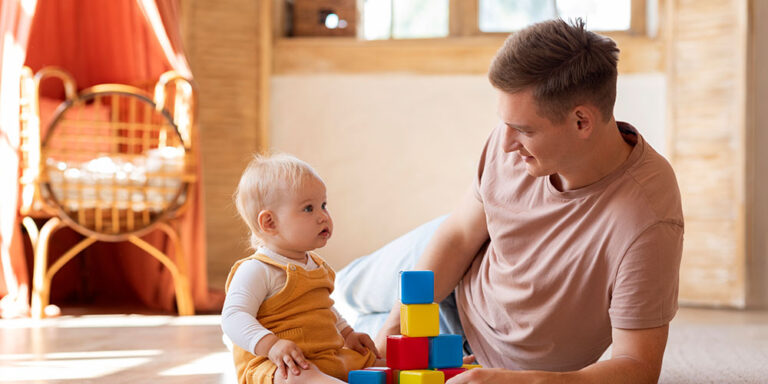 Vater spielt auf dem Boden mit seinem Baby, das bunte Bauklötze stapelt. Im Hintergrund steht ein Kinderbett.