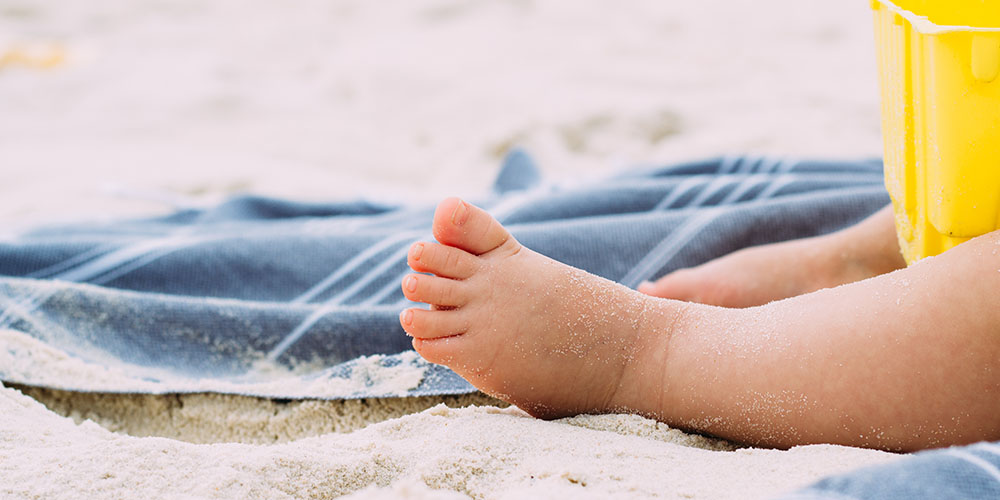 Nahaufnahme eines Babys, das am Strand auf einer blauen Decke sitzt, mit sandigen Füßen.