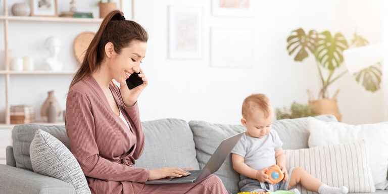 Junge Mutter arbeitet im Wohnzimmer an einem Laptop und telefoniert, während ihr Baby neben ihr auf dem Sofa sitzt und mit einem Spielzeug spielt.