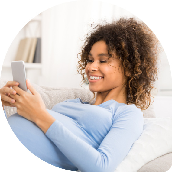 Eine schwangere Frau mit lockigem Haar sitzt entspannt auf einem Sofa und lächelt, während sie auf ihr Smartphone schaut. Sie trägt ein hellblaues Oberteil und ist von einer hellen, gemütlichen Umgebung umgeben. Die Atmosphäre wirkt ruhig und zufrieden.