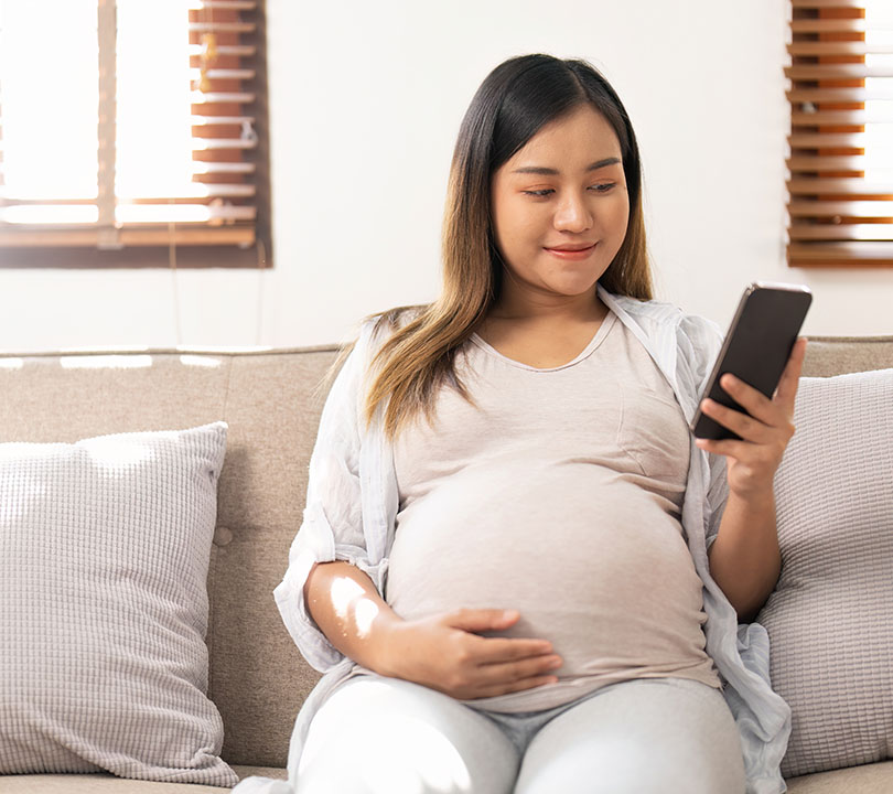 Junge schwangere Frau sitzt auf der Couch im Wohnzimmer und schaut zufrieden auf ihr Smartphone. Die Umgebung ist sehr hell und freundlich.