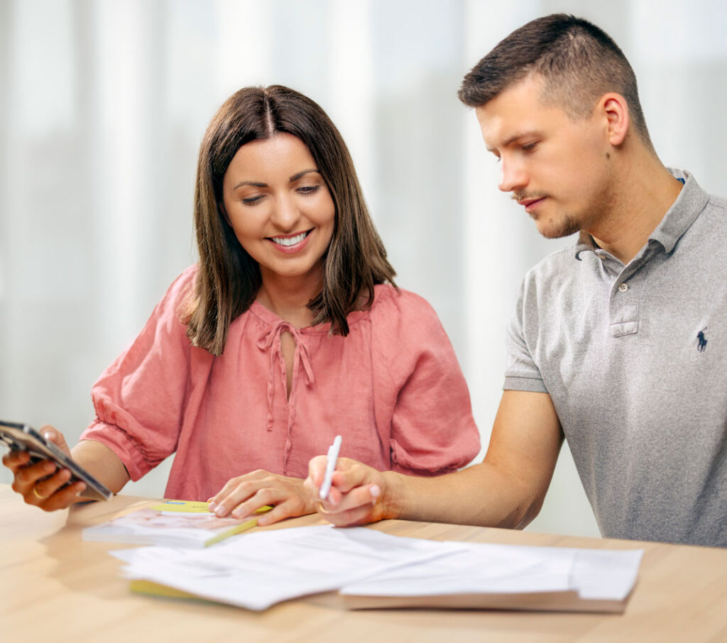 Paulina und Ingo Köpp, die Gründer von Elterngeld Buddy, sitzen an einem Tisch und arbeiten gemeinsam an Dokumenten. Paulina trägt eine rosafarbene Bluse und lächelt, während Ingo in einem grauen Poloshirt konzentriert etwas notiert. Die Szene ist in einem hellen Raum mit weichem Licht, was eine freundliche und produktive Atmosphäre schafft.