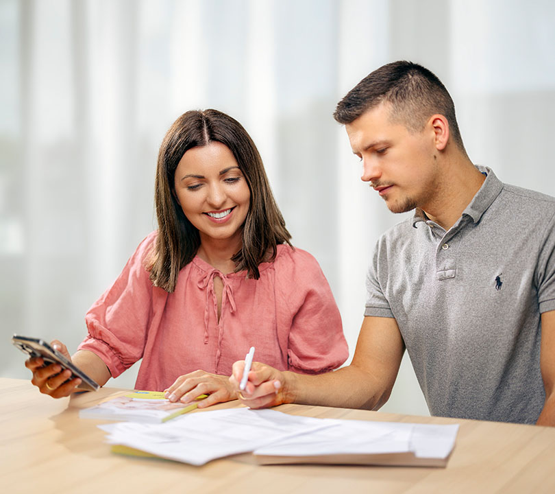 Paulina und Ingo Köpp, die Gründer von Elterngeld Buddy, sitzen an einem Tisch und arbeiten gemeinsam an Dokumenten. Paulina trägt eine rosafarbene Bluse und lächelt, während Ingo in einem grauen Poloshirt konzentriert etwas notiert. Die Szene ist in einem hellen Raum mit weichem Licht, was eine freundliche und produktive Atmosphäre schafft.